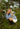 Woman squatting down in a grassy area outdoors, wearing a blue and white striped crewneck with a white skirt.