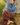 A woman sits outdoors against a tree trunk wearing a loose blue turtleneck sweater, brown wide-leg patterned pants, and brown leather loafers.