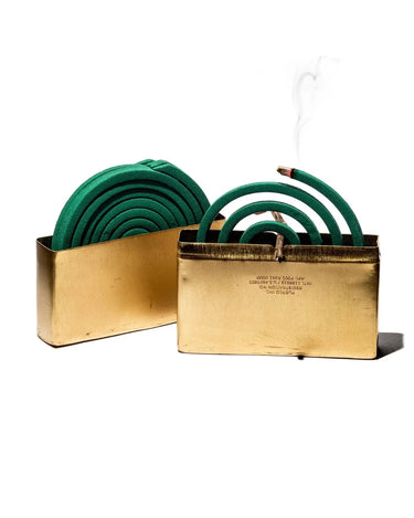 Gold mosquito coil holder on a white background, holding a green coil with visible smoke rising from the front burner.