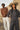 Two people standing in a desert landscape, one wearing a brown sweater and white pants, the other in a dark outfit with a wide-brimmed hat.