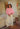 Woman standing in a room, wearing a pink crewneck sweater and white pants.
