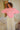 Woman standing in a room, wearing a pink crewneck sweater and white pants.