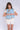 Woman standing in front of a white fabric backdrop wearing a light blue vest with brown trim over a white short sleeve top, paired with white bloomer-style shorts.