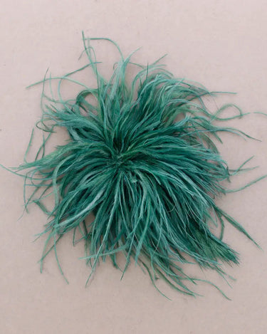 A fluffy, feathery green decorative brooch, photographed from directly above on a plain beige background.