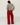 A woman shown from behind wearing a light gray knit sweater and bright red wide-leg pleated trousers, standing on a white wooden floor against a white wall.