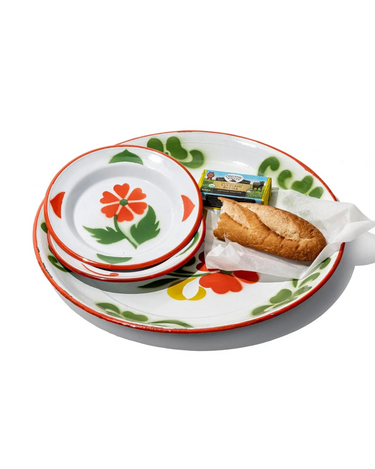A set of white ceramic plates with a colorful floral and leaf pattern, shown with a sandwich on white paper and a small packaged snack on a white background.