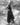 A woman stands in a grassy outdoor field wearing a long dark sleeveless dress and tall boots, photographed in black and white.