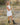 A woman stands in a grassy outdoor field at sunset wearing a sleeveless white dress and dark cowboy boots, holding a thin yellow flower.