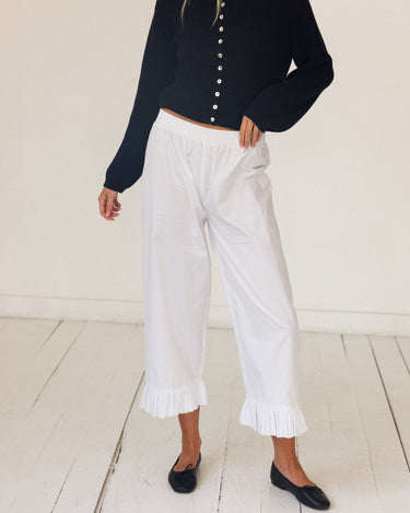 A model wearing white pants with ruffled hems, paired with a navy blue long-sleeve button-front cardigan and black flats, standing on a white wooden floor against a white wall.