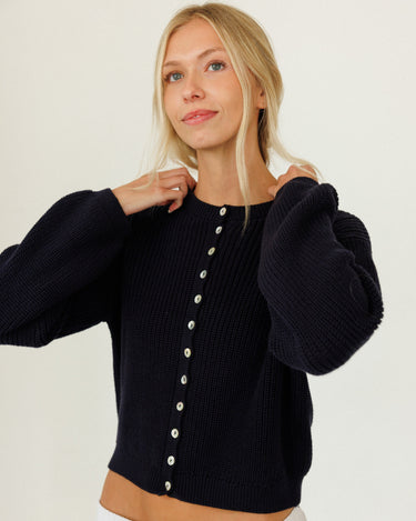 A woman wearing a black ribbed knit cropped sweater with balloon sleeves and a button-down front, paired with white pants, against a plain white background.