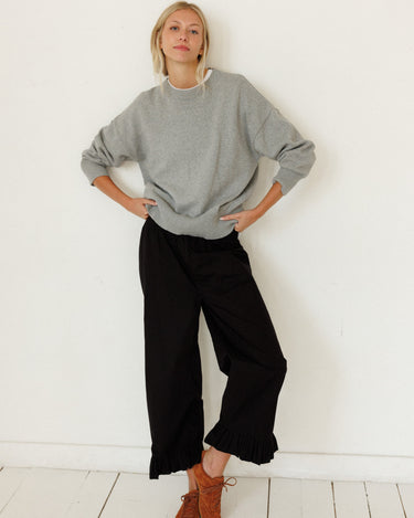 A woman posing in a light gray crewneck sweatshirt, black wide-leg cropped pants, and brown lace-up boots against a white wall and floor.