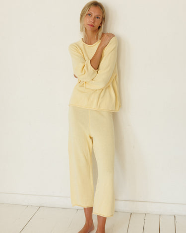 A woman wearing a pale yellow lounge set consisting of a long-sleeve sweater and matching pants, standing barefoot against a white wall on a white wooden floor.