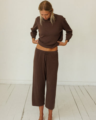 A woman wearing a matching brown lounge set consisting of a long-sleeve cropped top and wide-leg cropped pants, standing barefoot on a white wooden floor against a white wall.