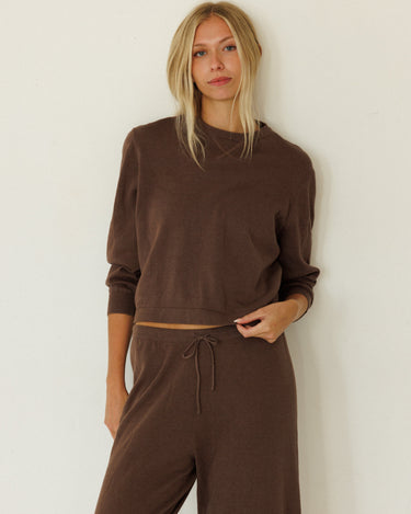 A woman modeling a matching brown lounge set consisting of a cropped long-sleeve sweatshirt and drawstring sweatpants, standing against a plain off-white wall.