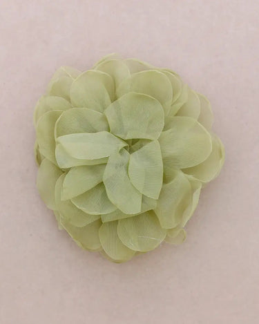 A single pale olive green fabric flower accessory, made of layered sheer petal-like ruffles, centered on a solid light pink background.