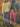 Two people standing on a leaf-covered ground in front of a wooden fence, one wearing blue gingham pants and a beige sweater, the other in red gingham pants and a denim jacket.