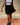 Close-up of a woman wearing a thin strap tank top and black skirt with black loafers against a beige background.