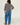 A woman is shown from behind wearing a navy blue and grey striped long-sleeve sweater and wide-leg blue jeans, standing in a bright white room with white wooden floorboards.
