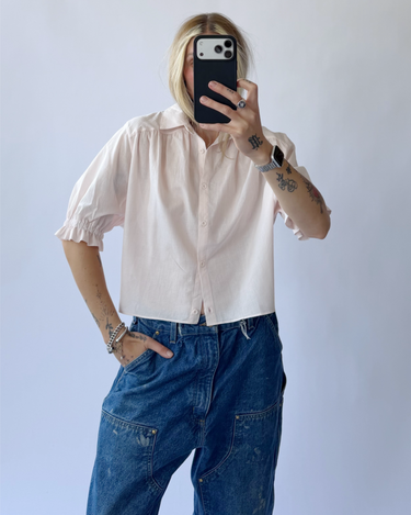 Person wearing a light pink shirt and blue jeans taking a mirror selfie.