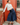 A woman wearing a white mock-neck long-sleeve top and a navy blue pleated midi skirt, leaning against a red brick door with a red metal railing.