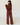 A woman wearing a matching dark red knit two-piece outfit with a cropped long-sleeve top and wide-leg pants, standing in profile against a white wall and light wood floor.
