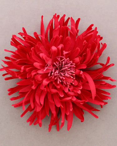A bright red fabric flower with layered, spiky petals arranged in a round daisy-like shape, photographed from above on a plain light gray background.