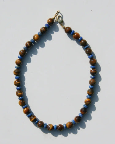 A beaded necklace made of brown tiger's eye beads and small blue spacer beads, with a gold clasp, laid flat on a light gray background.