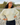 A woman wearing a pale green pleated quarter-sleeve top and a cream knit beanie, standing outdoors on a rocky path.
