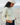 A woman wearing a pale green linen top and a striped beanie stands on a pebble beach, looking down.
