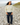 A woman stands on a rocky beach wearing a pale green quarter-sleeve cropped top, dark blue jeans, a cream striped beanie, and brown boots.