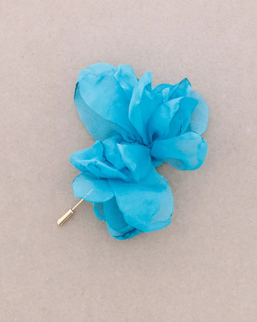A bright turquoise blue fabric flower brooch with a gold-tone safety pin clasp, displayed on a neutral beige background.