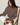 A woman sits on a wooden chair wearing a brown and white horizontally striped long-sleeve knit sweater, white pants, and brown clogs against a plain light background.