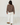 A woman shown from the back wearing a brown and white horizontally striped long-sleeve knit top, white wide-leg pants, and brown shoes, standing against a white wall and white wood floor.