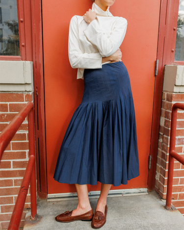 A person wearing a navy blue pleated midi skirt, a white long-sleeve mock-neck shirt, and brown leather loafers, standing in front of a red-orange brick door.