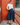 A woman stands outdoors against a red-orange door and brick wall, wearing a white mock-neck long-sleeve top, a dark navy blue pleated midi skirt, and brown leather loafers.