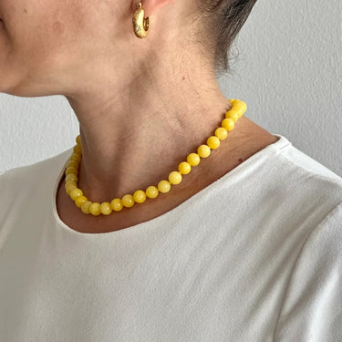Person wearing a yellow beaded necklace and gold hoop earrings against a plain background
