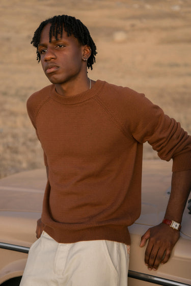 Man wearing a brown sweater and white pants standing next to a car with a blurred background