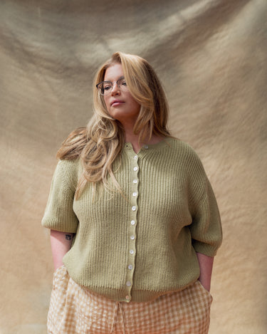 Woman wearing a green knit short sleeve cardigan and tan gingham pants in front of a tan fabric backdrop.