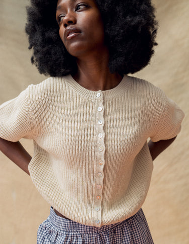 Woman wearing a beige knit short sleeve cardigan on a tan fabric backdrop.
