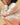 Close-up image of woman squatting down on a tan fabric backdrop wearing a cream, long-sleeve pointelle top and matching pointelle pants.