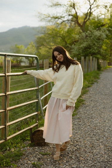 Woman wearing a yellow and white striped long-sleeved crewneck sweater over a pink skirt, standing in the outdoors holding a fence.