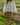 Woman holding a wooden basket in a grassy field outdoors wearing a white eyelet long sleeve blouse and tan skirt.