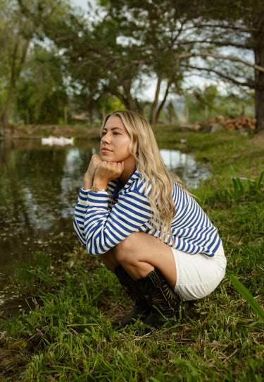 Woman squatting down in a grassy area outdoors, wearing a blue and white striped crewneck with a white skirt.