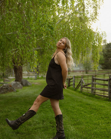Woman in a grassy area outdoors wearing a short, black slip dress with thin spaghetti straps and black cowboy boots.
