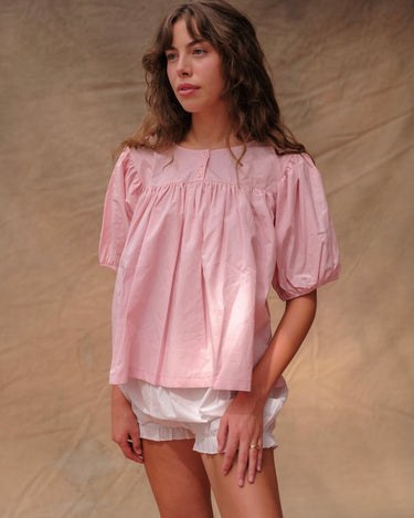 Woman wearing a pink, short-sleeved babydoll-style blouse and white bloomer shorts on a tan fabric background.