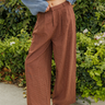 A person wearing brown wide-leg pleated trousers with a small check pattern, styled with a blue knit sweater and brown leather loafers, standing outdoors in front of a green hedge.