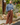 A woman stands outdoors on a sidewalk wearing a blue-gray turtleneck sweater and brown wide-leg trousers, with brown loafers, in front of a wooden utility pole and garden plants.