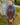 A woman sits on grass in front of a tree trunk, wearing a blue mock-neck sweater, brown plaid print pants, and brown loafers.