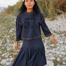 A woman wearing a dark navy blue t-shirt with a large black '57' graphic on the front, gold contrast stitching, and a white striped knit beanie, standing outdoors against a blurred natural background.