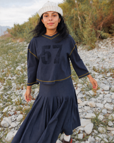 A woman wearing a dark navy blue t-shirt with a large black '57' graphic on the front, gold contrast stitching, and a white striped knit beanie, standing outdoors against a blurred natural background.
